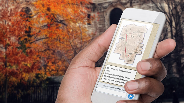 A hand holding a phone playing Treasure Hunt Manchester with a tree with orange leaves behind.
