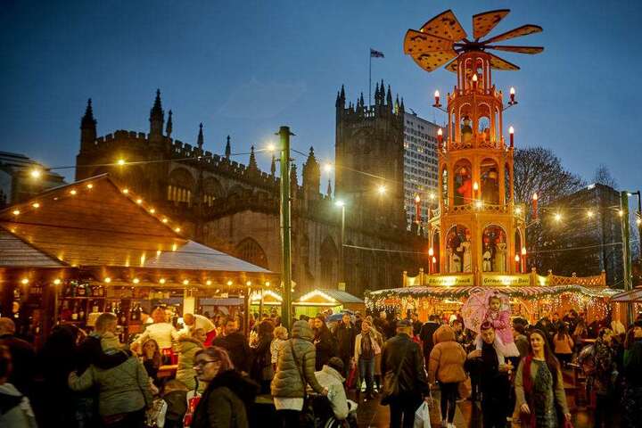 Manchester Christmas market in twilight with a crowd of visitors, festive stalls, a towering, illuminated windmill decoration, and Manchester Cathedral the background.